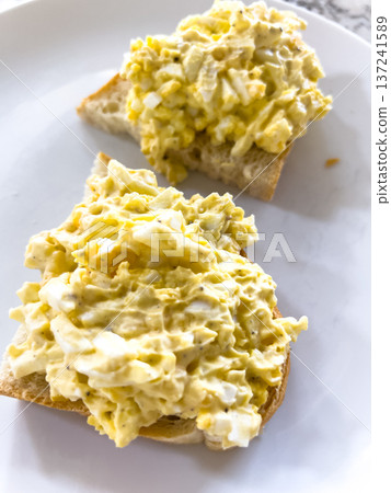 Two open-faced slices of toasted bread topped with homemade egg salad placed on a white kitchen plate. Two open-faced slices of toasted bread topped with homemade egg salad placed on a white kitchen plate. 137241589