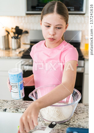 A girl in a pink shirt prepares dry ingredients in a glass bowl, holding a container of baking powder. The kitchen background is bright and modern. A girl in a pink shirt prepares dry ingredients in a glass bowl, holding a container of baking powder. The kitchen background is bright and modern. 137241616