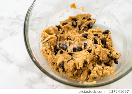 A glass bowl holds chocolate chip cookie dough on a white marble surface. Overhead lighting highlights the thick and textured dough. 137241627