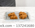 Gingerbread cookies, including a gingerbread man with a heart-shaped cutout, rest on a rustic metal tray against a marble countertop. 137241680