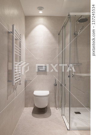 Modern bathroom with a glass shower, white toilet, and towel radiator. Neutral color scheme with tiled walls and floor. Clean and minimalist design. Modern bathroom with a glass shower, white toilet, and towel radiator. Neutral color scheme with tiled walls and floor. Clean and minimalist design. 137245534