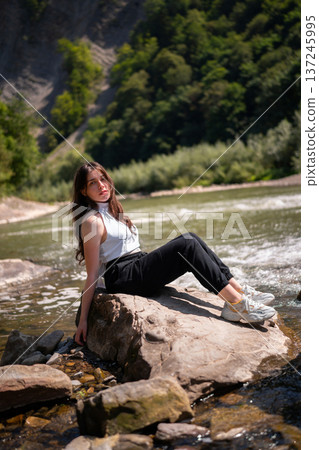 Relaxed Young Woman Resting on Rocks by a Mountain River 137245995