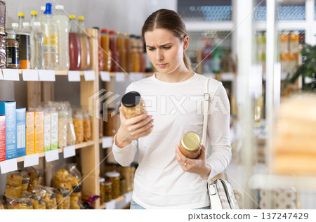 Girl customer in supermarket examines jar of kidney bean and lentil, chooses food-stuff in grocery Girl customer in supermarket examines jar of kidney bean and lentil, chooses food-stuff in grocery 137247429