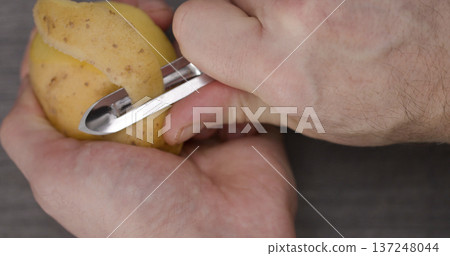 Person peels a potato with a peeler in a kitchen setting during the daytime preparing for cooking 137248044