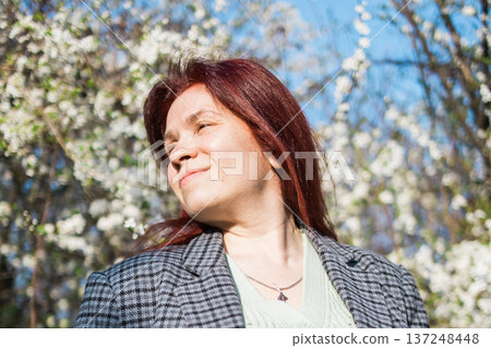 Woman enjoying sunlight under blooming trees in spring garden. Seasonal renewal, wellbeing, outdoor relaxation, nature connection, flowering branches, warm daylight, peaceful lifestyle during spring 137248448