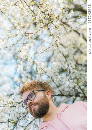 Man standing under flowering tree branches during bright spring bloom. Contemplation, calm lifestyle, connection with nature, seasonal renewal and peaceful outdoor moment in blooming spring garden Man standing under flowering tree branches during bright spring bloom. Contemplation, calm lifestyle, connection with nature, seasonal renewal and peaceful outdoor moment in blooming spring garden 137248698