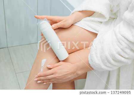 Close up hands of woman in a white bathrobe holding a white lotion bottle and applying cream to her legs, performing a daily skincare routine for moisturized and healthy skin Close up hands of woman in a white bathrobe holding a white lotion bottle and applying cream to her legs, performing a daily skincare routine for moisturized and healthy skin 137249095