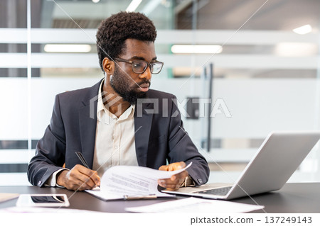 Concentrated professional man reviewing documents and working on a laptop at a modern office desk, managing business affairs and planning new strategies Concentrated professional man reviewing documents and working on a laptop at a modern office desk, managing business affairs and planning new strategies 137249143