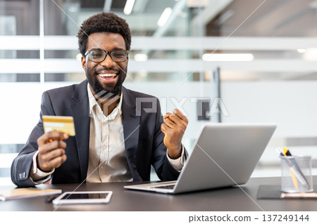 Happy man smiling widely, holding a golden credit card and making a triumphant fist gesture, actively engaging in online shopping or banking on a laptop in a modern office Happy man smiling widely, holding a golden credit card and making a triumphant fist gesture, actively engaging in online shopping or banking on a laptop in a modern office 137249144