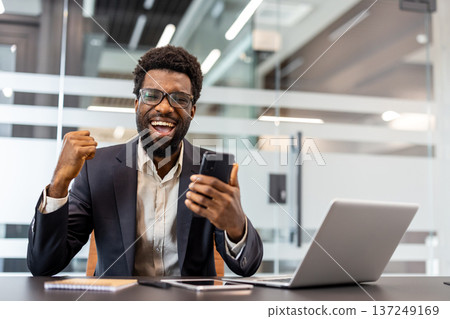 Businessman expressing excitement and confidence while celebrating victory at work, happily receiving good news and positive results on a mobile phone in a modern office setup Businessman expressing excitement and confidence while celebrating victory at work, happily receiving good news and positive results on a mobile phone in a modern office setup 137249169