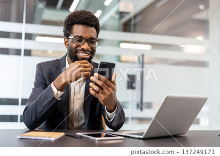 Smiling businessman browsing smartphone at a desk with open laptop in a modern office, looking positive and connected while working remotely and staying productive and professional 137249171