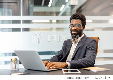 Young professional man wearing eyeglasses typing on a laptop, looking at camera and smiling while sitting at his desk in a contemporary corporate environment Young professional man wearing eyeglasses typing on a laptop, looking at camera and smiling while sitting at his desk in a contemporary corporate environment 137249186