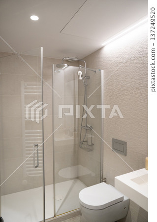 Modern bathroom with frameless glass shower, rain and handheld heads, floating toilet and integrated white sink countertop against textured beige tiles and recessed lighting Modern bathroom with frameless glass shower, rain and handheld heads, floating toilet and integrated white sink countertop against textured beige tiles and recessed lighting 137249520