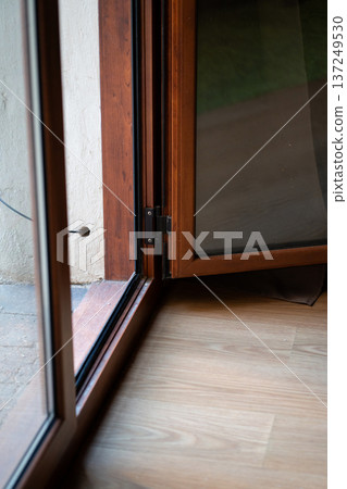 Close-up of open brown wooden PVC balcony door frame and glass pane featuring a visible hinge mechanism, connecting indoor wooden floor with outdoor concrete patio Close-up of open brown wooden PVC balcony door frame and glass pane featuring a visible hinge mechanism, connecting indoor wooden floor with outdoor concrete patio 137249530