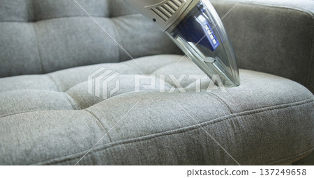 Vacuum cleaner removes dust from a gray couch in a bright living room during the day 137249658