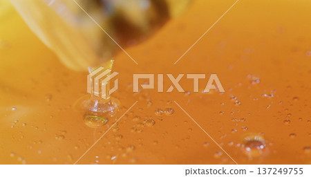 Honey drips into a bowl of bright orange liquid, creating bubbles and ripples in the surface 137249755