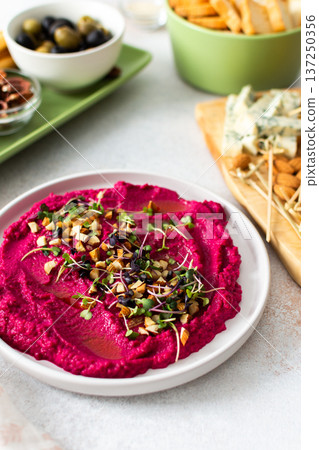 Beetroot hummus is placed in the center of a charcuterie board, surrounded by snacks like olives, nuts, cheese, and crispy breadsticks. A light, inviting setup showcases vibrant colors Beetroot hummus is placed in the center of a charcuterie board, surrounded by snacks like olives, nuts, cheese, and crispy breadsticks. A light, inviting setup showcases vibrant colors 137250356