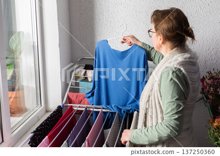 A woman around 50 years old is hanging clothes on a drying rack outside her balcony. She carefully organizes items to let them dry in the sunlight during the afternoon A woman around 50 years old is hanging clothes on a drying rack outside her balcony. She carefully organizes items to let them dry in the sunlight during the afternoon 137250360