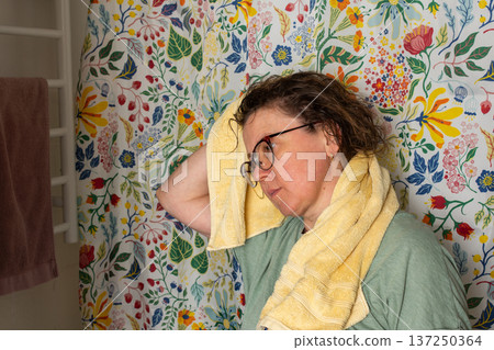 A 50-year-old woman starts her morning routine in the bathroom. She looks at herself in the mirror while holding a towel over her shoulder. The colorful background adds brightness A 50-year-old woman starts her morning routine in the bathroom. She looks at herself in the mirror while holding a towel over her shoulder. The colorful background adds brightness 137250364