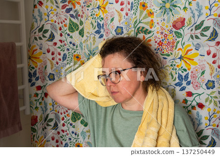 A 50-year-old woman starts her morning routine in the bathroom. She looks at herself in the mirror while holding a towel over her shoulder. The colorful background adds brightness 137250449