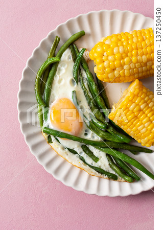 A plate holds fresh asparagus, a fried egg with a yolk, and sweet corn on the cob. The setting features a soft pink background with a simple fork beside the food 137250450
