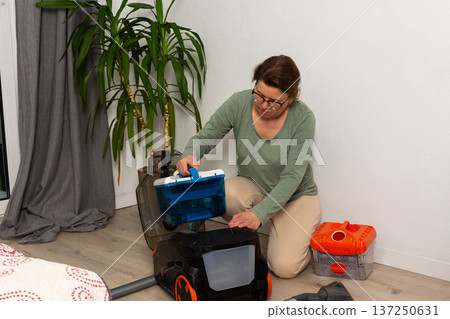 In a living room, a person is getting a vacuum cleaner ready for cleaning. The person is seated on the floor with the vacuum cleaner open and looks focused on the task In a living room, a person is getting a vacuum cleaner ready for cleaning. The person is seated on the floor with the vacuum cleaner open and looks focused on the task 137250631
