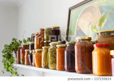 Canned vegetables and fruits are lined up on a shelf in a kitchen, showing winter preparations. A painting of a basket is seen in the background as part of the decor 137250654