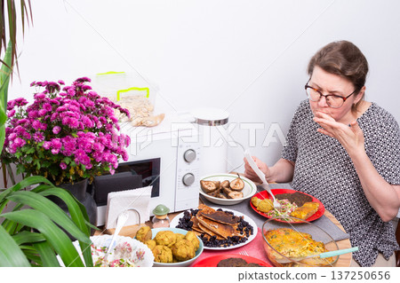 A person sits at a kitchen table eating food. Dishes with different homemade meals are arranged around her. Flowers add color to the table setting. A microwave is nearby 137250656