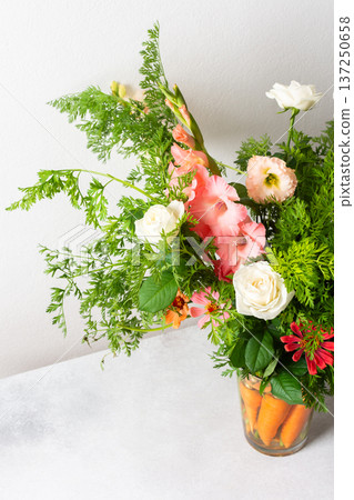 A bouquet of roses, gladioli, zinnias, and eustomas stands in water. Carrots with green tops are part of the arrangement on a plain table against a light background A bouquet of roses, gladioli, zinnias, and eustomas stands in water. Carrots with green tops are part of the arrangement on a plain table against a light background 137250658