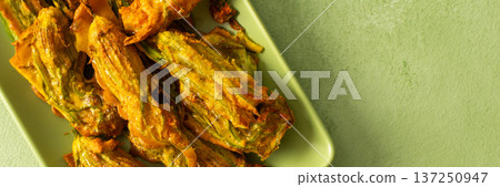 Crispy, golden-brown deep-fried zucchini flowers sit on a light green plate. They are perfect for sharing at a summer gathering, offering a delicious flavor and unique texture, banner 137250947