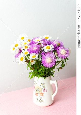 A colorful bouquet of daisies and purple flowers arranged in a charming pitcher rests on a pink tablecloth, bringing joy to the soft, simple setting A colorful bouquet of daisies and purple flowers arranged in a charming pitcher rests on a pink tablecloth, bringing joy to the soft, simple setting 137251082