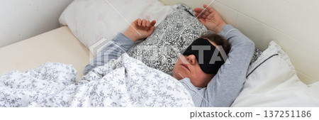 A 50-year-old woman is sound asleep in her bed, wearing a sleep mask. She is surrounded by pillows and blankets, creating a tranquil atmosphere. Sunlight gently filters into the room, banner A 50-year-old woman is sound asleep in her bed, wearing a sleep mask. She is surrounded by pillows and blankets, creating a tranquil atmosphere. Sunlight gently filters into the room, banner 137251186