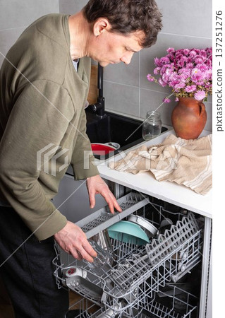 A man is putting dirty dishes into the dishwasher in his kitchen. He focuses on the task while several items rest in the appliance, ready for cleaning A man is putting dirty dishes into the dishwasher in his kitchen. He focuses on the task while several items rest in the appliance, ready for cleaning 137251276