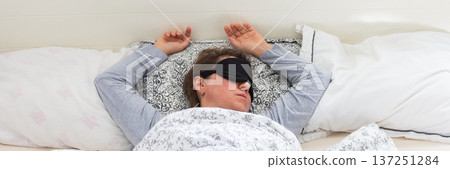 A 50-year-old woman is comfortably sleeping in her bed with a sleep mask on. She is wrapped in a soft blanket, resting peacefully in a serene bedroom setting, banner A 50-year-old woman is comfortably sleeping in her bed with a sleep mask on. She is wrapped in a soft blanket, resting peacefully in a serene bedroom setting, banner 137251284