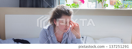 A tired 50-year-old woman sits on her bed, rubbing her eyes as she adjusts to the early morning light. Her cozy bedroom features plants and flowers visible outside the window, banner A tired 50-year-old woman sits on her bed, rubbing her eyes as she adjusts to the early morning light. Her cozy bedroom features plants and flowers visible outside the window, banner 137251291