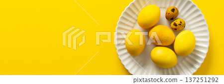 A decorative plate holds several yellow Easter eggs, symbolizing spring and renewal, alongside a couple of speckled quail eggs, all against a vibrant yellow background, banner 137251292