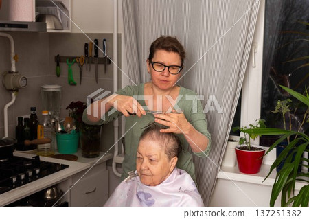 A woman is cutting her mother hair in the kitchen while they enjoy time together. The setting features plants and flowers, creating a lively atmosphere for this home activity A woman is cutting her mother hair in the kitchen while they enjoy time together. The setting features plants and flowers, creating a lively atmosphere for this home activity 137251382