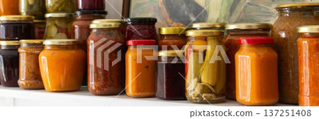 Canned vegetables and fruits are lined up on a shelf in a kitchen, showing winter preparations. A painting of a basket is seen in the background as part of the decor, banner 137251408