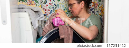 Woman stands in bathroom putting clothes into washing machine while surrounded by towels and laundry, carrying out daily household task with focus and care, banner Woman stands in bathroom putting clothes into washing machine while surrounded by towels and laundry, carrying out daily household task with focus and care, banner 137251412