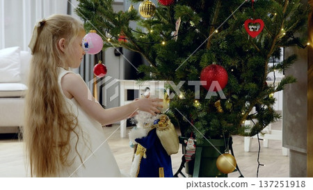 Young girl wearing white dress hanging colorful ornaments on christmas tree, positioning santa claus figurine while creating festive holiday atmosphere at home Young girl wearing white dress hanging colorful ornaments on christmas tree, positioning santa claus figurine while creating festive holiday atmosphere at home 137251918