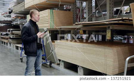 Man utilizing smartphone in lumber warehouse, examining wood panels for diy project. Numerous options in the aisles. shelves with lumber. translator: good price Man utilizing smartphone in lumber warehouse, examining wood panels for diy project. Numerous options in the aisles. shelves with lumber. translator: good price 137251921