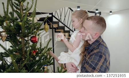 Smiling father helping young daughter hanging ornaments on christmas tree, creating festive moment in warmly lit home living room during holiday season Smiling father helping young daughter hanging ornaments on christmas tree, creating festive moment in warmly lit home living room during holiday season 137251935