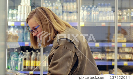Young woman carefully choosing food products from refrigerated shelves in a supermarket, reading labels and making purchasing decisions Young woman carefully choosing food products from refrigerated shelves in a supermarket, reading labels and making purchasing decisions 137252024