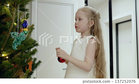 Blond girl carefully hanging glittery red christmas ornament while decorating festive tree inside contemporary living space during seasonal celebration 137252076