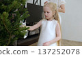 Young girl decorating christmas tree with owl ornament while mother tenderly brushing her long blonde hair, capturing warm holiday family scene 137252078