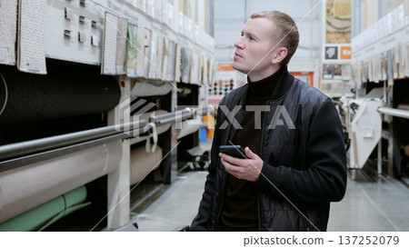 Focused interior designer comparing carpets on smartphone in warehouse, making decisions for renovation project. Professional entrepreneur using technology for online shopping 137252079