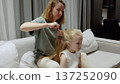 Mother gently braiding daughter's long blond hair, sharing intimate moment on living room sofa, depicting loving family connection and nurturing relationship 137252090