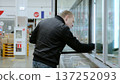 Man carefully opens a freezer door in a busy supermarket, selecting frozen food items with concentration. Cold air fills the aisle as he browses, capturing a typical shopping experience 137252093