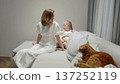 Loving mother wearing white dress sitting on cozy sofa with young daughter, ginger cat napping peacefully nearby, sharing tender family moment 137252119