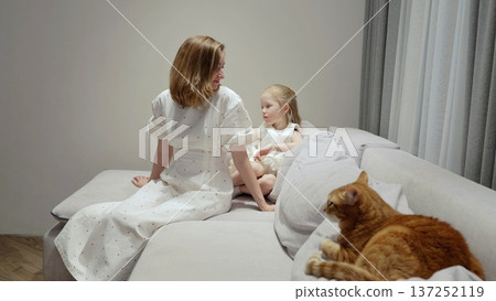 Loving mother wearing white dress sitting on cozy sofa with young daughter, ginger cat napping peacefully nearby, sharing tender family moment 137252119